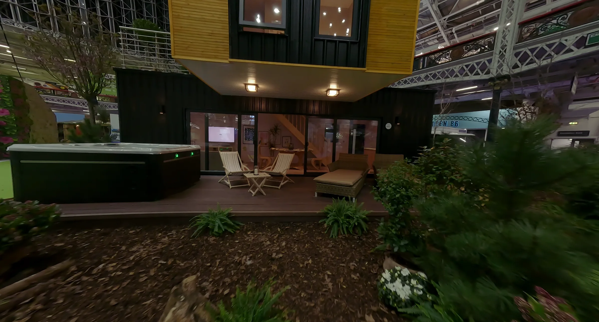 Front of the two-storey exhibition build: timber-clad upper floor cantilevered over the glazed ground floor, terrace and hot tub