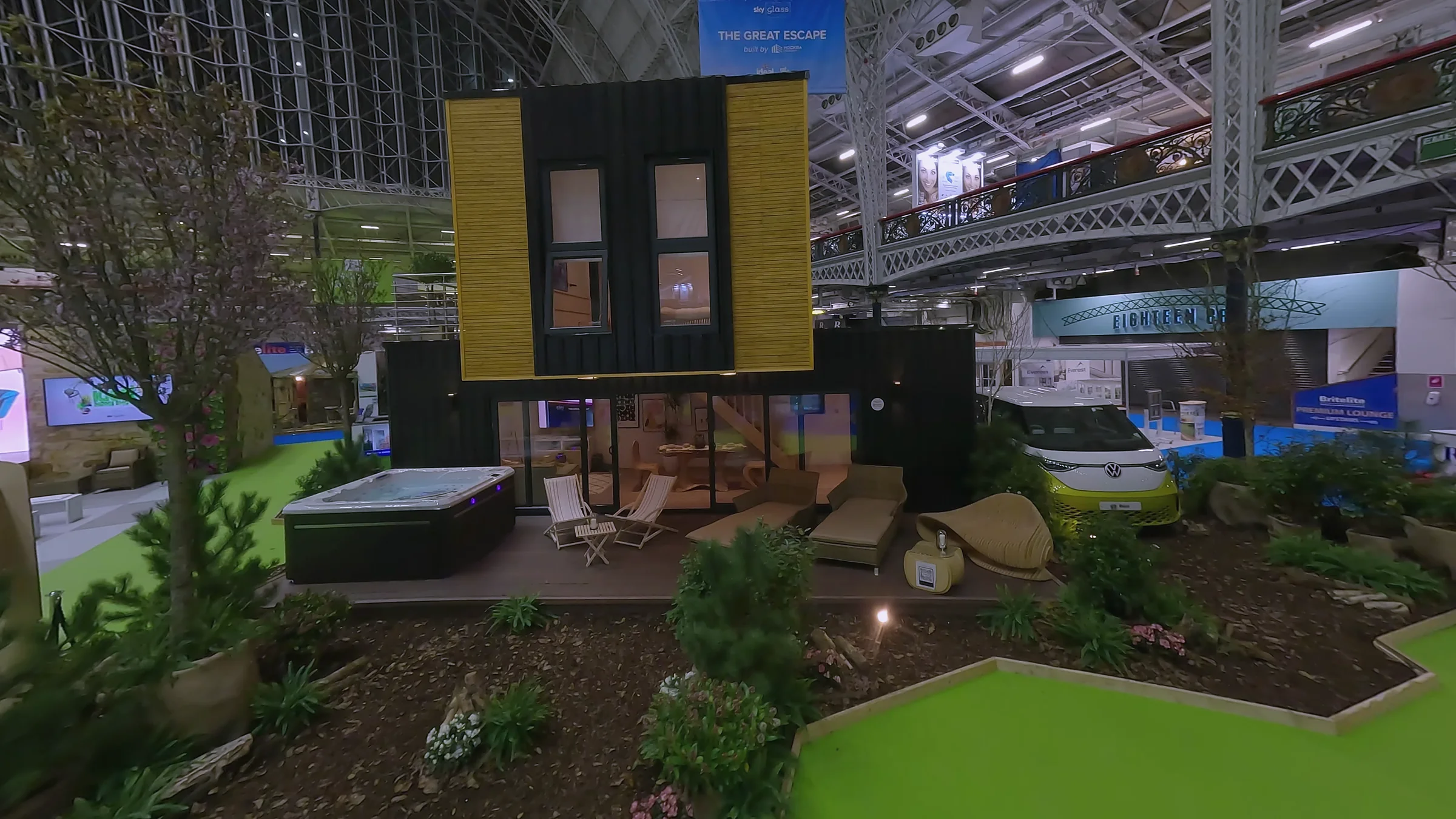 Two-storey timber-clad exhibition build with cantilevered upper floor, terrace, hot tub and planted garden, exhibited inside the Ideal Home Show hall
